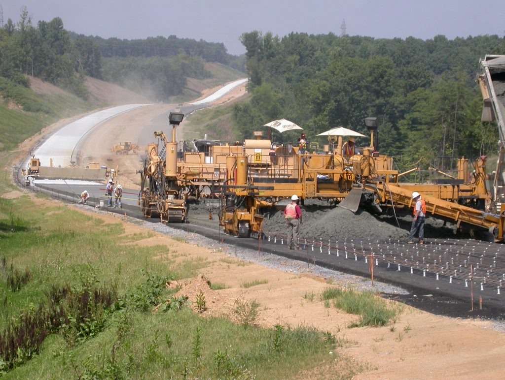 Image: Photograph shows a construction crew working on US 29 near Lynchburg, Virginia in 2004. The image shows workers and equipment laying and smoothing the base in the foreground; the new concrete roadway of the bypass is shown in the background on hilly terrain with trees on either side of the roadway.